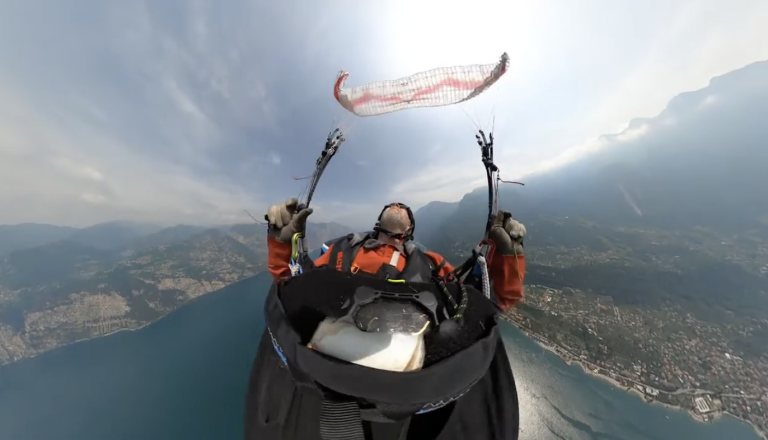 Paraglider pilot in an orange jacket, seen from a 360 camera, holding the brakes with the wing stalled overhead during SIV training above Lake Garda.